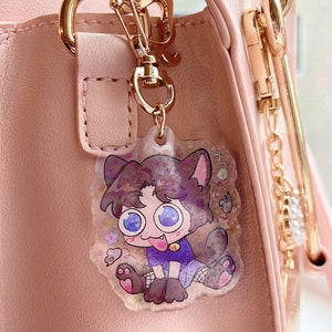 May include: A clear acrylic charm featuring a cartoon cat-like character with big blue eyes, a purple outfit, and a lolling tongue. The charm is attached to a gold-colored keychain and is hanging on a pink purse.