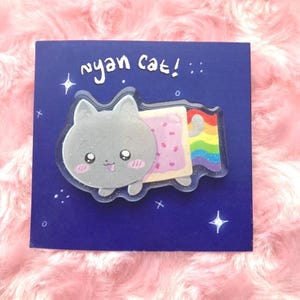 May include: A Nyan Cat pin on a dark blue card. The pin features a gray cat with a pink-cheeked face, a Pop-Tart body, and a rainbow trail. The words "nyan cat!" are written in white above the pin, with star accents.
