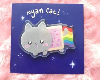 Pode incluir: Um alfinete Nyan Cat em um cartão azul escuro. O alfinete apresenta um gato cinza com rosto de bochechas rosadas, corpo de Pop-Tart e rastro de arco-íris. As palavras "nyan cat!" estão escritas em branco acima do alfinete, com estrelas.