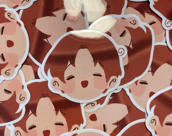 May include: A pile of stickers depicting a cartoon face with closed eyes, a small open mouth, and rosy cheeks. The hair is brown, and the face is a light peach colour. The stickers have a white border and a glossy finish.