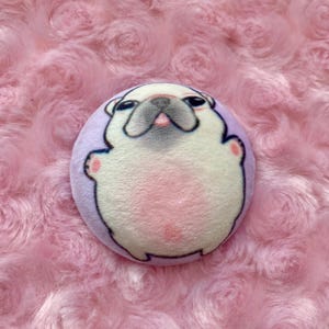 Pode incluir: Um broche redondo de pelúcia com um pug. O pug tem um corpo branco, barriga rosa e contorno preto. O rosto do pug tem um nariz preto, olhos e uma língua rosa. O broche está sobre um fundo rosa texturizado.