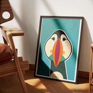 May include: Framed art print featuring a stylized illustration of a puffin bird. The artwork has a teal background with the puffin in black, white, orange, and beige. The print is in a black frame and leans against a wall.