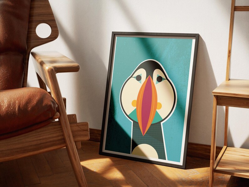 May include: Framed art print featuring a stylized illustration of a puffin bird. The artwork has a teal background with the puffin in black, white, orange, and beige. The print is in a black frame and leans against a wall.