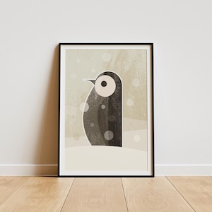 May include: A framed art print of a stylized penguin illustration. The penguin is in shades of grey and black, set against a snowy backdrop with falling white dots. The artwork is framed in a black border and rests on a wooden floor.