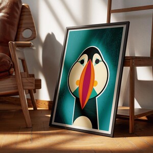 May include: Framed art print featuring a stylized illustration of a puffin bird. The artwork has a teal background with the puffin's face in cream, black, and orange. The bird's beak is a vibrant combination of red and orange.