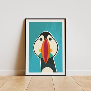 May include: Framed art print featuring a stylized illustration of a puffin bird. The artwork has a teal background with a geometric design of the bird's face, showcasing a large orange beak, black and white head, and teal accents. The print is in a black frame.