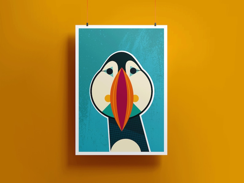 May include: A framed art print featuring a stylized illustration of a puffin. The bird has a black head, white cheeks, and a large, colorful beak with orange, red, and teal accents. The background is a textured teal color.