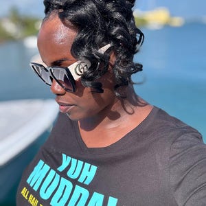 May include: A woman wearing black sunglasses with a white frame and a black t-shirt with the text "Yuh Mudda! All Hail The Virgin Islands" in blue and yellow lettering.