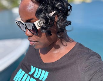 Può includere: Una donna indossa occhiali da sole neri con montatura bianca e una maglietta nera con la scritta "Yuh Mudda! All Hail The Virgin Islands" in lettere blu e gialle.