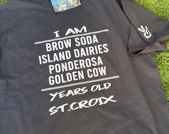 May include: Black t-shirt with white text that reads "I am Brow Soda Island Dairies Ponderosa Golden Cow Years Old St. Croix".
