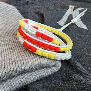 May include: A stack of colorful bracelets with red, yellow, and white beads. The bracelets are resting on a gray knit hat and a dark gray fabric with a white design.