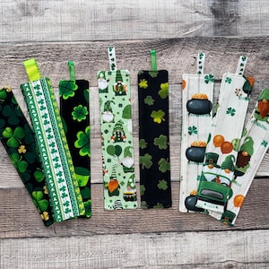 May include: A collection of St. Patrick's Day themed fabric bookmarks. Designs include shamrocks, gnomes, and pots of gold. Each bookmark has a fabric loop at the top. The bookmarks are arranged on a wooden surface.