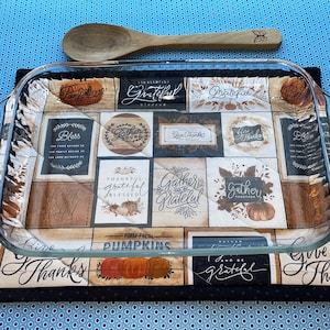 May include: A black and brown quilted hot pad with a Thanksgiving theme. The pad features a variety of designs, including pumpkins, leaves, and the words "Gather" and "Give Thanks". A glass baking dish is sitting on top of the hot pad.