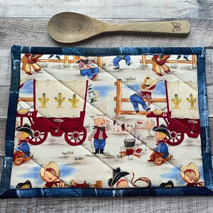 May include: A quilted rectangular hot pad with a denim blue border and a cowboy-themed print. The print features cowboys, covered wagons, cacti, and a campfire scene. A wooden spoon with a butterfly design rests above.