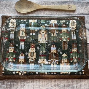 May include: A glass baking dish sits on a brown quilted hot pad with a pattern of nutcracker soldiers on a green background. The hot pad is on a wood surface.