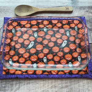 May include: A purple and black Halloween-themed hot pad with a glass baking dish on top. The hot pad features a repeating pattern of orange pumpkins with black eyes and a white ghost.