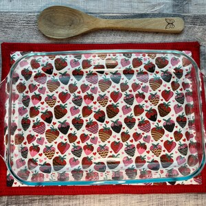 May include: A red and white fabric hot pad with a glass baking dish on top. The hot pad has a pattern of strawberries with chocolate and pink accents.
