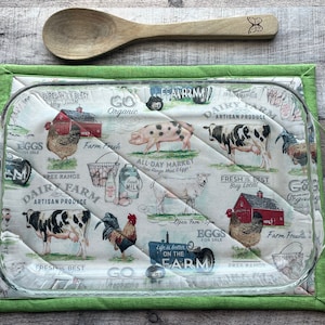 May include: A green quilted hot pad with a farmhouse design featuring a rooster, pig, cows, and a red barn. The text on the pad reads "Go Organic", "Dairy Farm Artisan Produce", "Fresh is Best Buy Local", and "Eggs on the Farm".