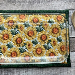 May include: A green and yellow quilted hot pad with a sunflower pattern. A clear glass baking dish is sitting on top of the hot pad. A wooden spoon with a butterfly design is on the right side of the image.