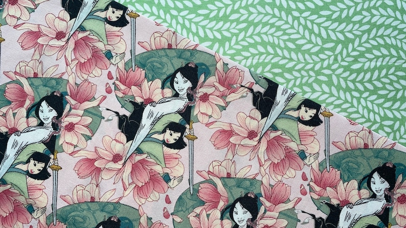 May include: Fabric featuring a repeating pattern of a woman in a traditional outfit, swords, and pink flowers on a light pink background. A section of green fabric with a white leaf pattern is in the upper right corner.