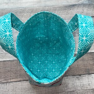 May include: A handmade tote bag with a turquoise and white floral pattern on the handles and interior lining. The bag's exterior features a subtle pattern, and the handles are arched. The bag is open, revealing its spacious interior.