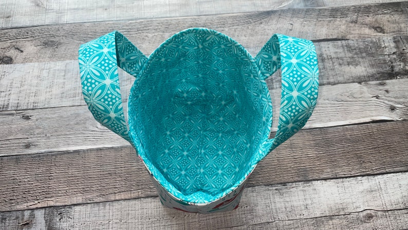 May include: A handmade tote bag with a turquoise and white floral pattern on the handles and interior lining. The bag's exterior features a subtle pattern, and the handles are arched. The bag is open, revealing its spacious interior.