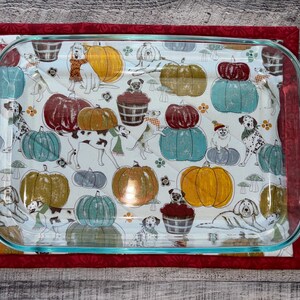 May include: A red quilted hot pad with a glass baking dish on top. The baking dish has a white background with a pattern of dogs, pumpkins, and fall leaves.