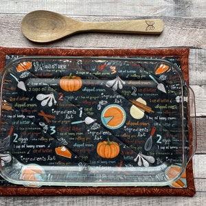 May include: A clear glass baking dish rests on a quilted, rectangular hot pad with a brown border. The pad features a black background with a pie recipe design, including pumpkins and text. A wooden spoon with a butterfly design is above.