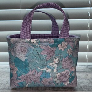May include: A small tote bag featuring a floral pattern with a cartoon princess design in shades of teal, lavender, and white. The handles are a solid purple with a chevron pattern. The interior lining is a solid purple.