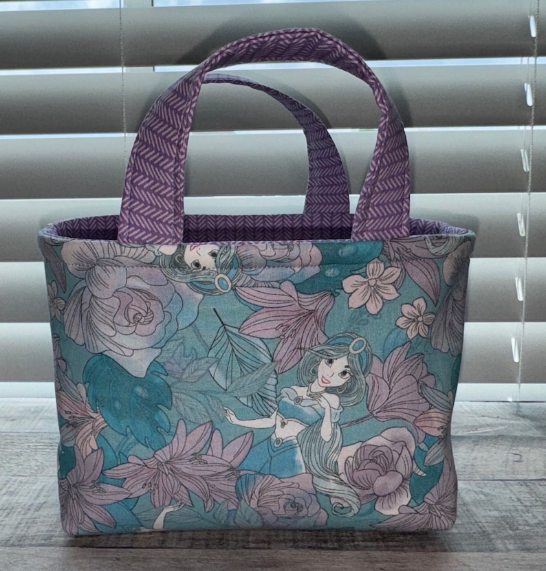 May include: A small tote bag featuring a floral pattern with a cartoon princess design in shades of teal, lavender, and white. The handles are a solid purple with a chevron pattern. The interior lining is a solid purple.