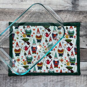 May include: A glass baking dish on top of a green and white quilted hot pad with a pattern of gnomes gardening.