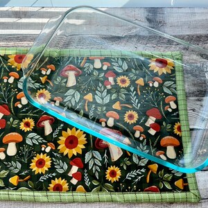 May include: A glass baking dish with a green and brown floral print hot pad. The hot pad features a repeating pattern of mushrooms, sunflowers, and leaves.