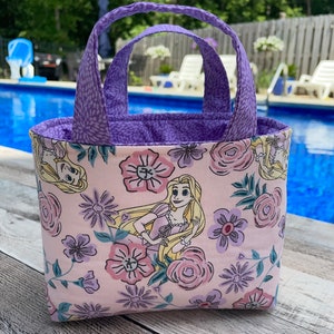 May include: A small, rectangular tote bag with a floral and character print. The bag features a light pink background with purple flowers and a cartoon character. The handles and interior lining are purple.