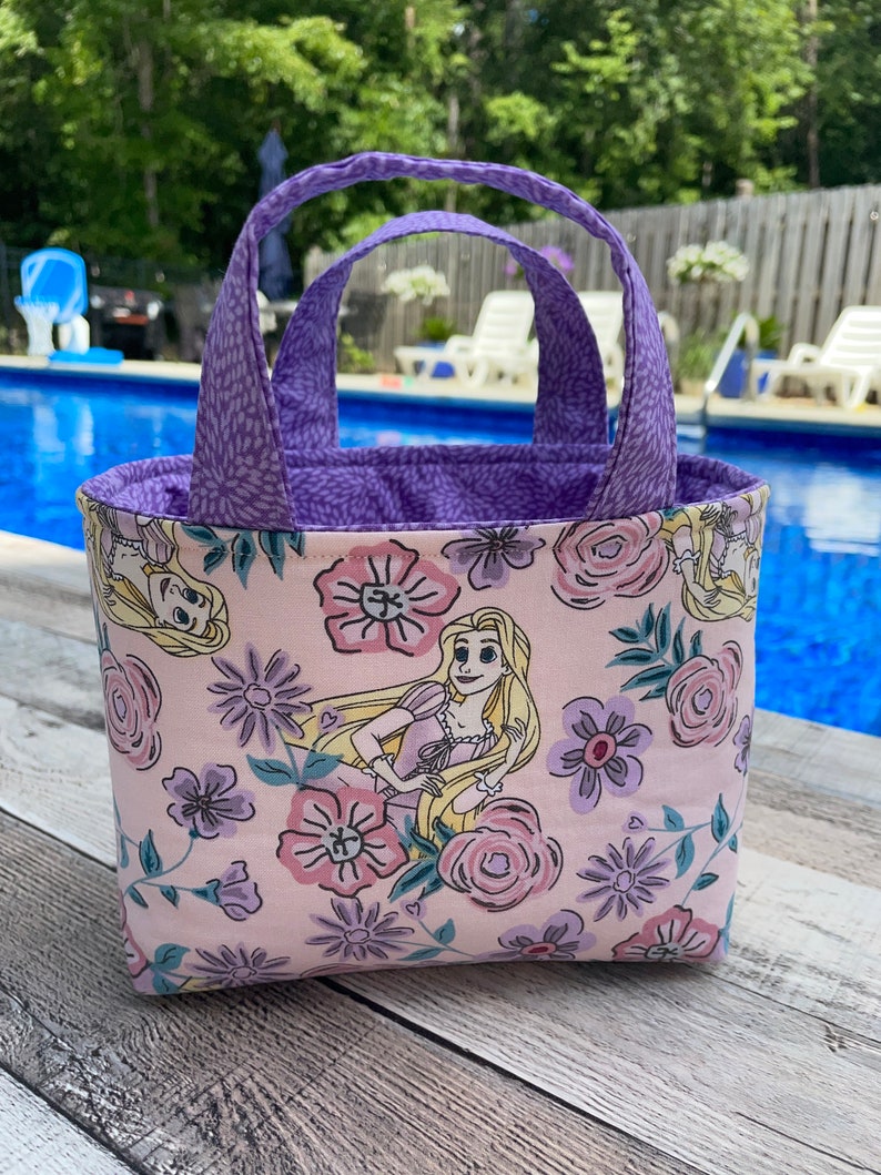 May include: A small, rectangular tote bag with a floral and character print. The bag features a light pink background with purple flowers and a cartoon character. The handles and interior lining are purple.