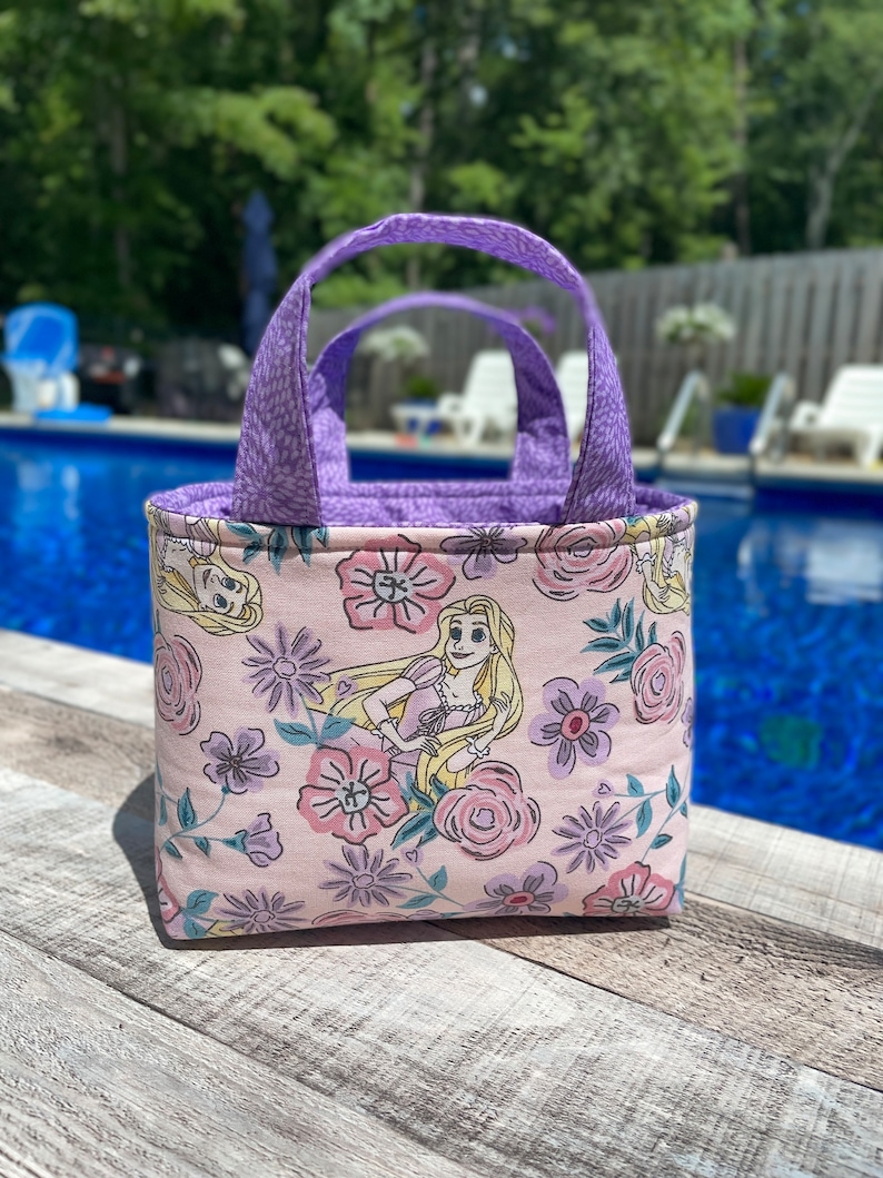 May include: A small, rectangular tote bag with a floral and character print. The bag features a light pink background with a pattern of flowers and a cartoon character. The handles and interior lining are a solid purple color.