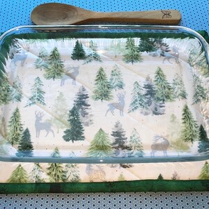 May include: A glass baking dish with a green and white woodland print featuring trees and deer. The dish is sitting on a green and white quilted hot pad.
