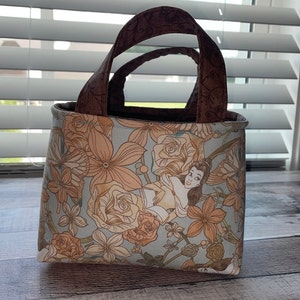 May include: A small tote bag with a floral pattern in shades of peach and light blue. The bag features a cartoon illustration of a woman. The handles are brown with a floral design. The bag is sitting on a wooden surface.