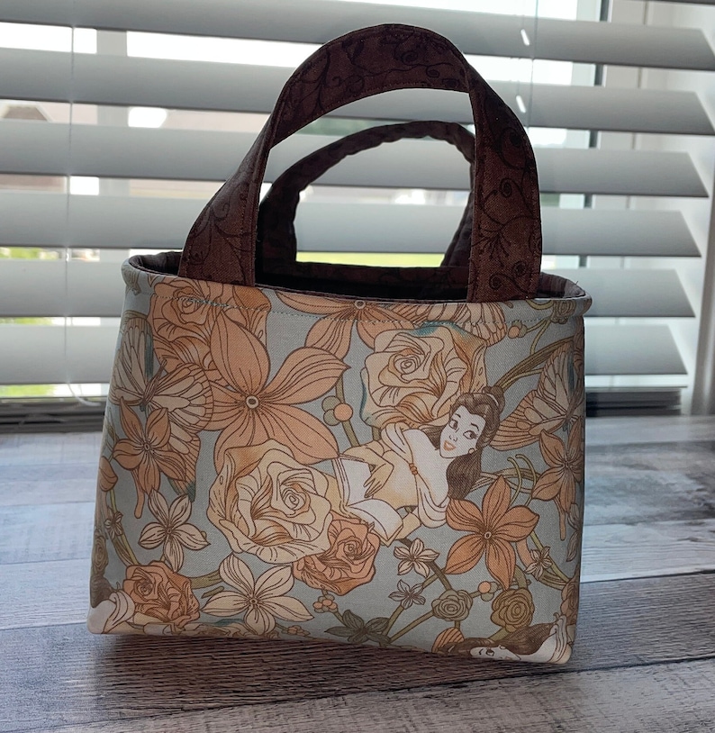 May include: A small tote bag with a floral pattern in shades of peach and light blue. The bag features a cartoon illustration of a woman. The handles are brown with a floral design. The bag is sitting on a wooden surface.