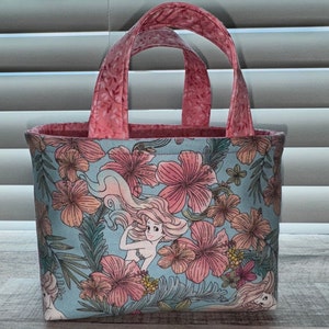 May include: A small tote bag with a tropical floral pattern in pink, peach, and green on a light blue background. The bag features a mermaid illustration and pink handles and interior lining.