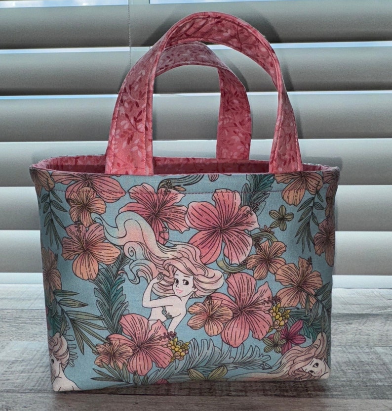 May include: A small tote bag with a tropical floral pattern in pink, peach, and green on a light blue background. The bag features a mermaid illustration and pink handles and interior lining.