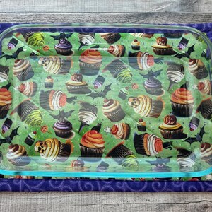 May include: A glass baking dish with a green and purple fabric hot pad. The fabric has a pattern of cupcakes with bats and other Halloween-themed designs.