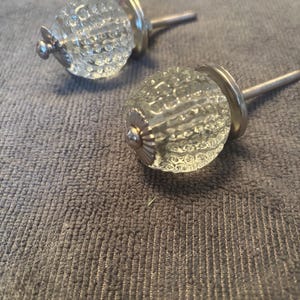 May include: Two clear glass cabinet knobs with a spherical, textured surface featuring a repeating dimpled pattern. Each knob has a metallic silver-toned base and a long, threaded metal bolt extending from the back for installation.