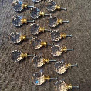 May include: A collection of fourteen clear, multifaceted glass cabinet knobs, each featuring a polished gold-tone metal base and a threaded metal screw extending from the back, arranged in a loose formation on a dark gray fabric surface.