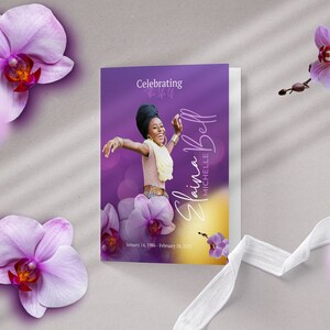 Op de afbeelding: Een paarse en gouden herdenkingskaart met een foto van een lachende vrouw op een witte achtergrond. De tekst luidt "Celebrating the Life of Elaina Michelle Bell, January 14, 1984 - February 18, 2023".