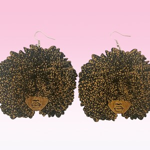 May include: A pair of wooden earrings featuring a black silhouette of a woman with an afro hairstyle. The earrings are brown and have a silver hook.