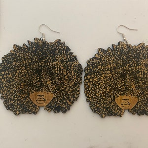 May include: A pair of wooden earrings featuring a black silhouette of a woman with an afro hairstyle. The earrings are brown and have silver hooks.