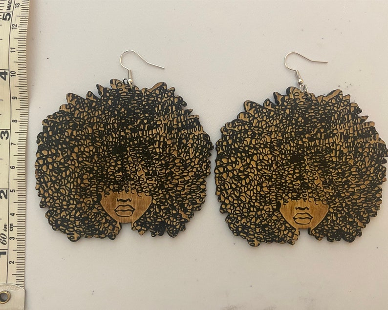 May include: A pair of wooden earrings featuring a black silhouette of a woman with an afro hairstyle. The earrings are brown and have silver hooks.