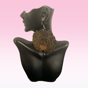 May include: A pair of black and gold earrings shaped like an afro. The earrings are made of fabric and have a wooden face in the center.