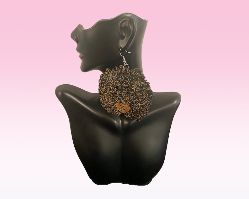 May include: A pair of black and gold earrings shaped like an afro. The earrings are made of fabric and have a wooden face in the center.