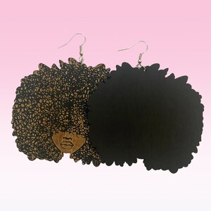 May include: A pair of wooden earrings featuring a black silhouette of a woman with an afro hairstyle. One earring has a detailed design of the woman's face and hair, while the other is a solid black afro.